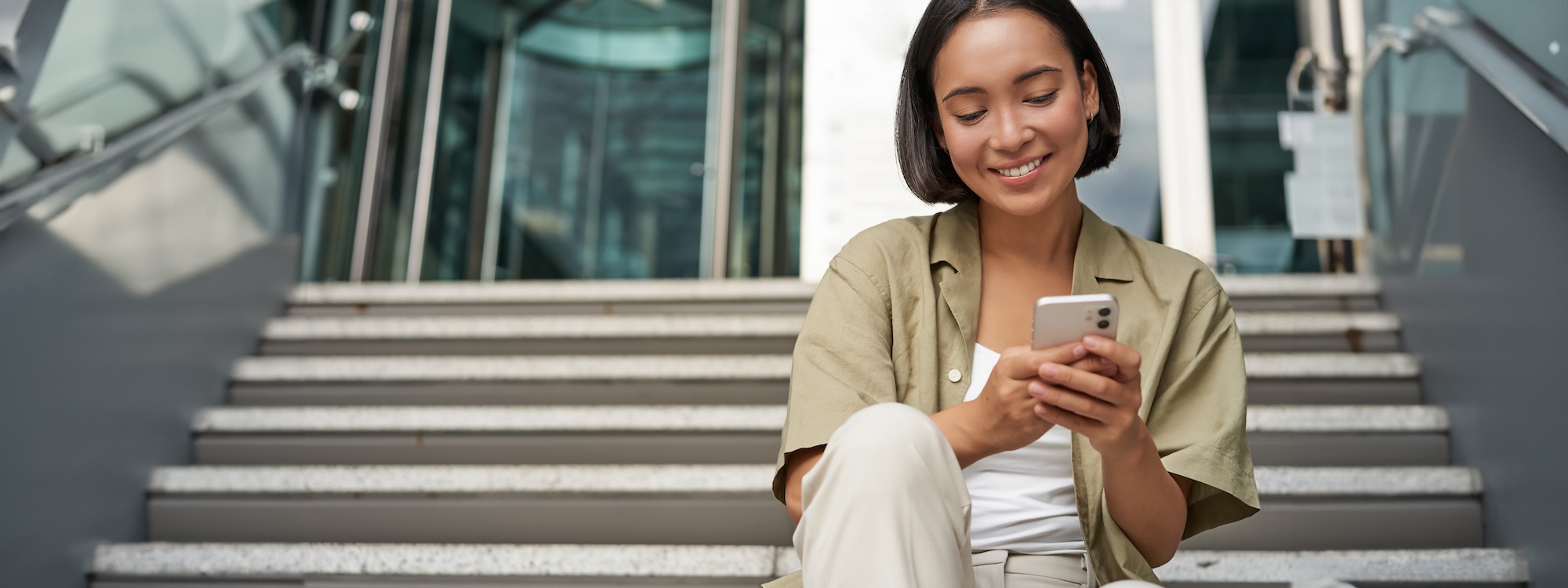 Smiling asian girl sits on stairs near building entrance, using mobile phone app. Happy young woman rests with smartphone in her hands outdoors